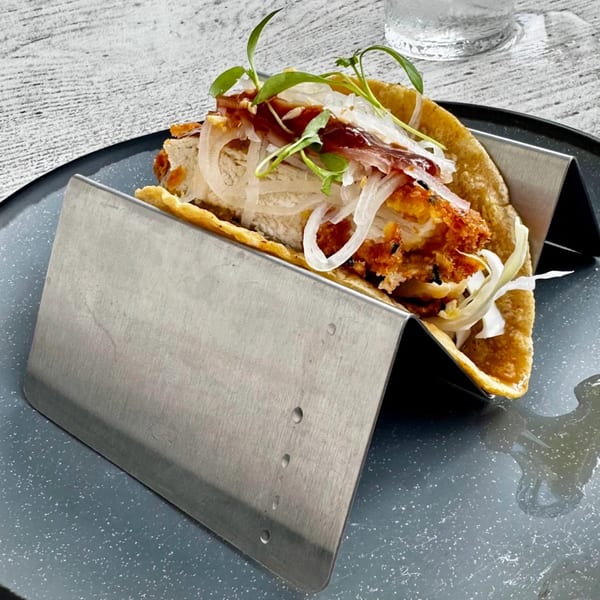 Crispy fried chicken taco with sliced onions, sauce, and microgreens in a metal holder.