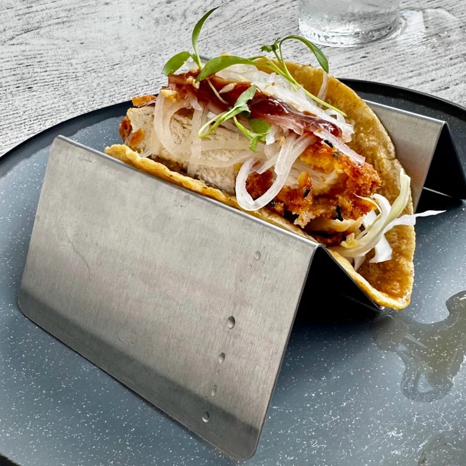 Crispy fried chicken taco with sliced onions, sauce, and microgreens in a metal holder.