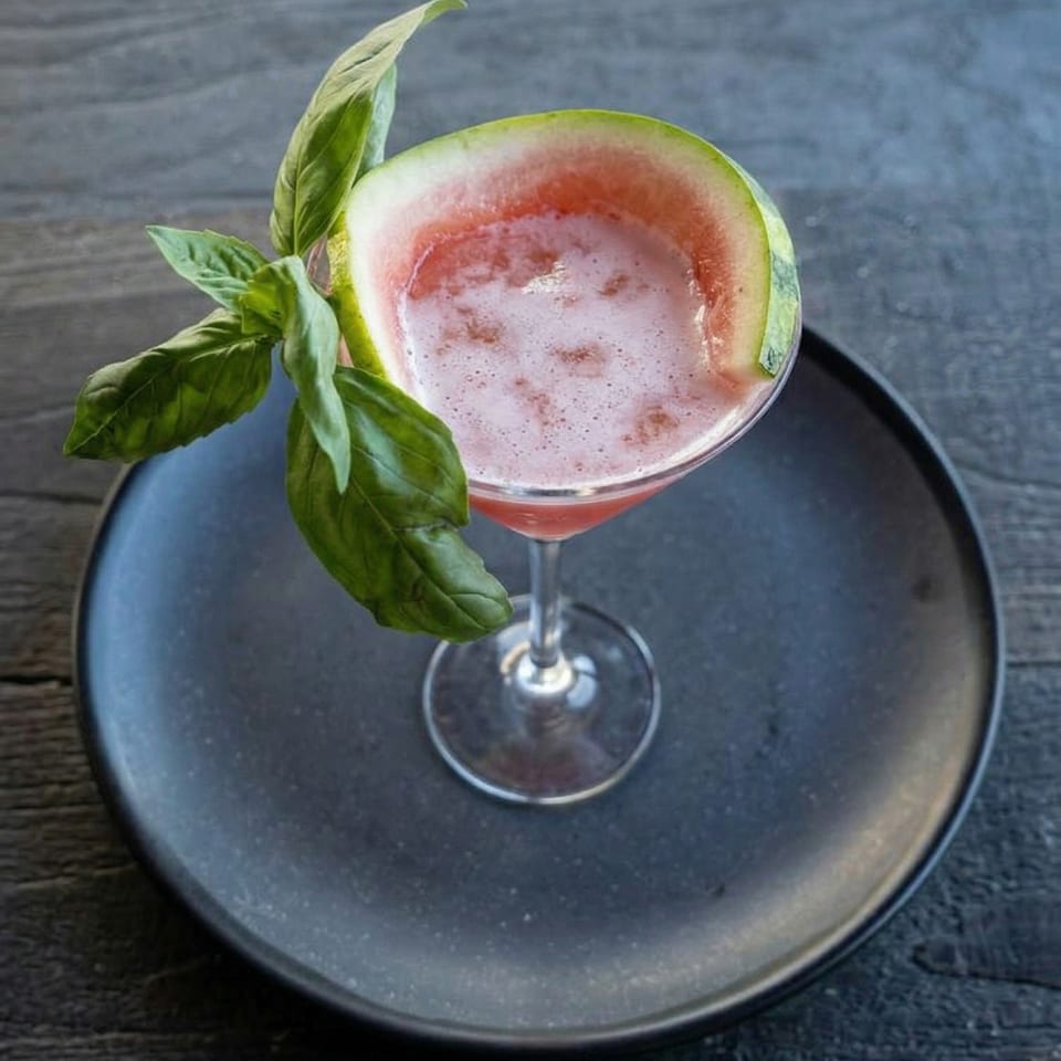 Watermelon cocktail with basil garnish and a watermelon slice, served on a dark plate.