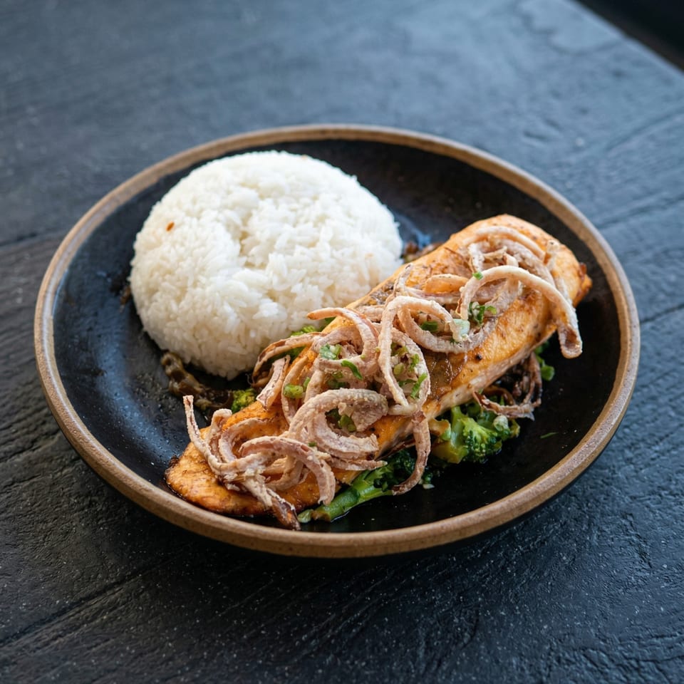 Salmon fillet topped with crispy fried onions, served with white rice and broccoli on a dark plate.