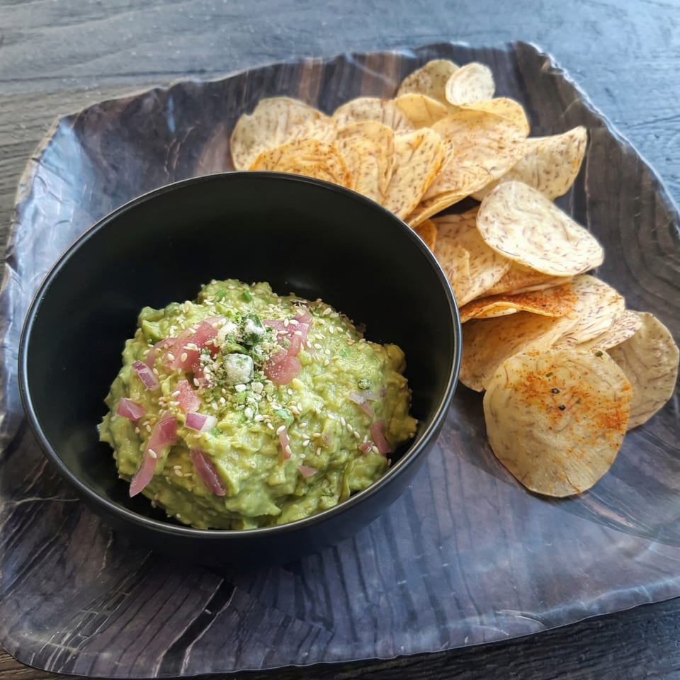 Guacamole topped with red onions and sesame seeds, served with taro chips on a dark plate.