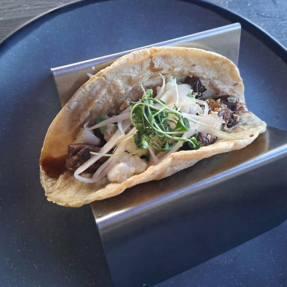 A single taco with meat, white onions, and green sprouts, served on a metal holder.