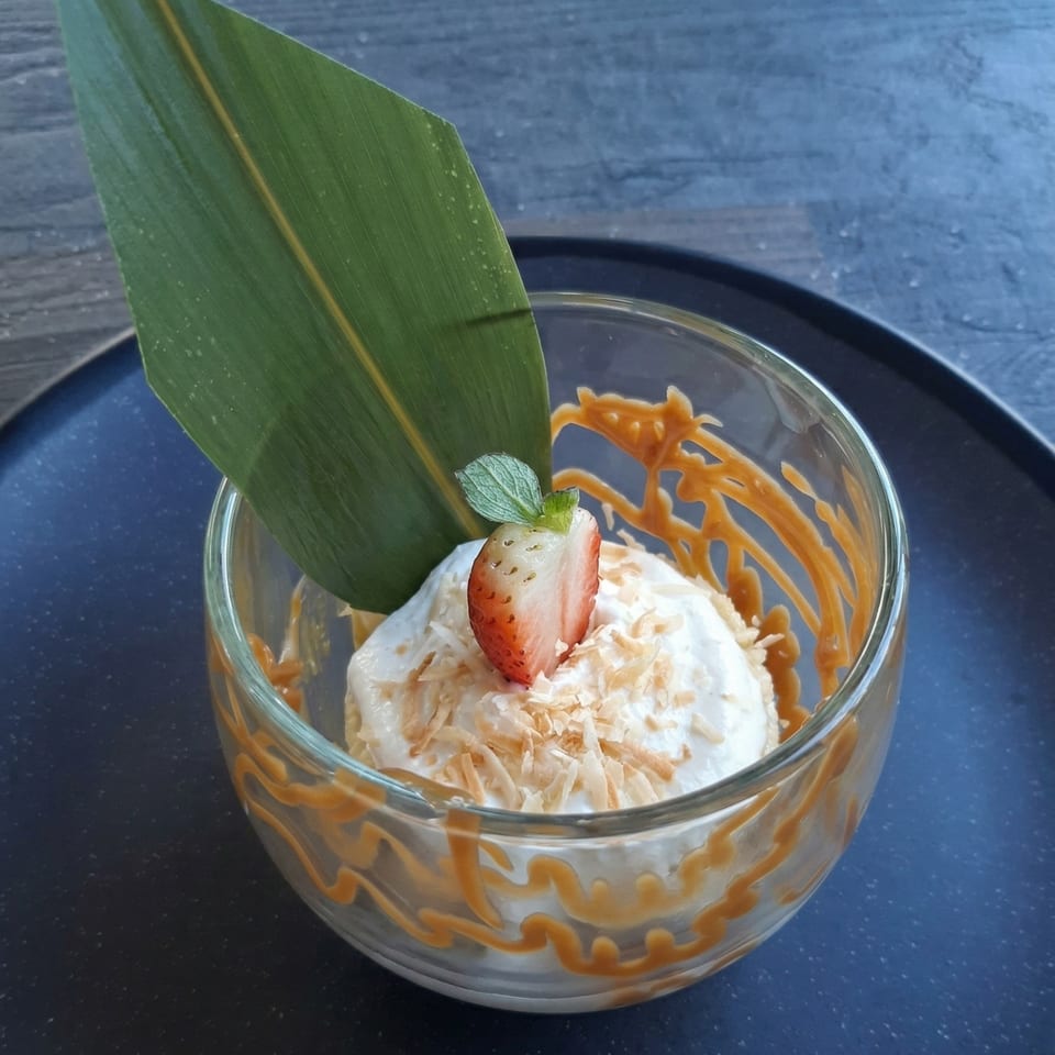 A dessert in a glass bowl with white cream, coconut flakes, a strawberry, and a green leaf.