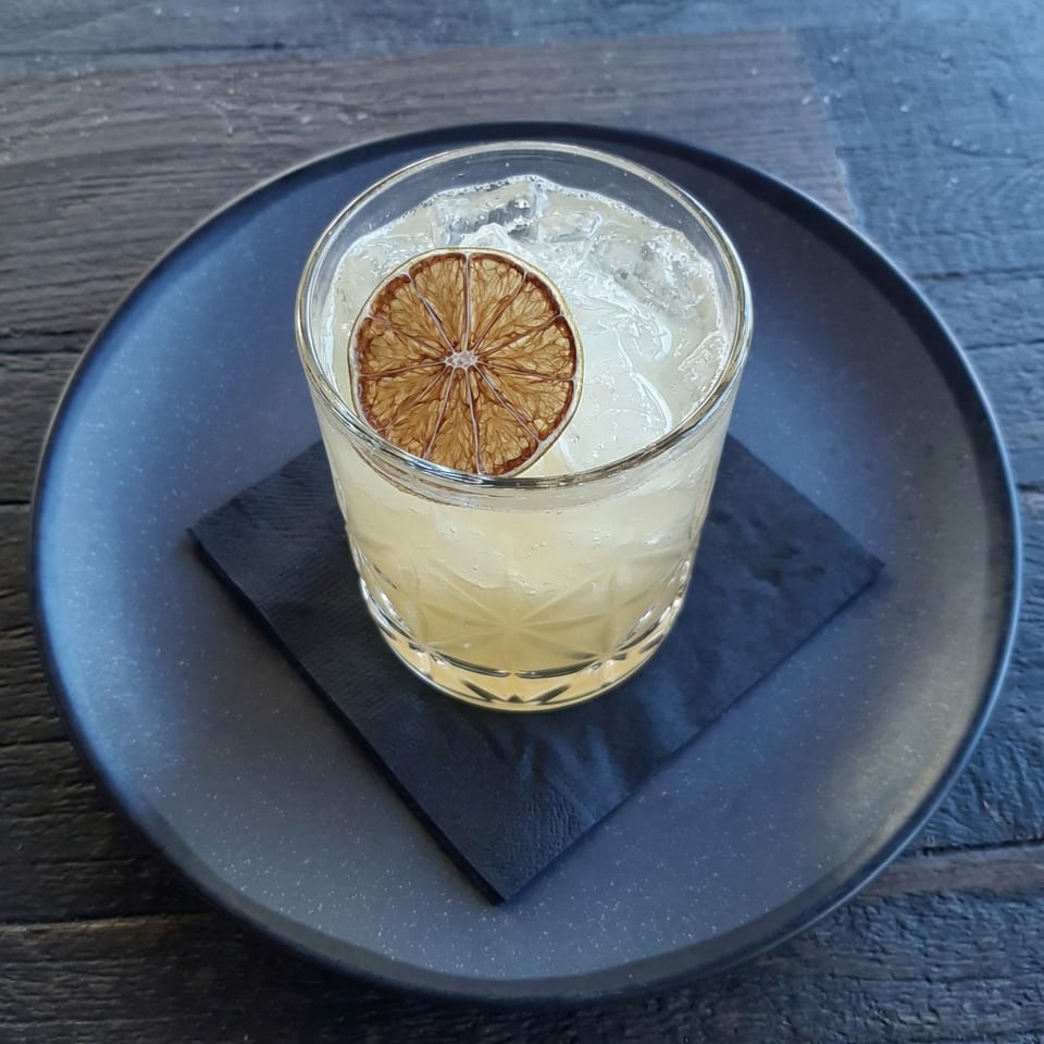 A light-colored cocktail with ice and a dried lime slice, on a black napkin and plate.