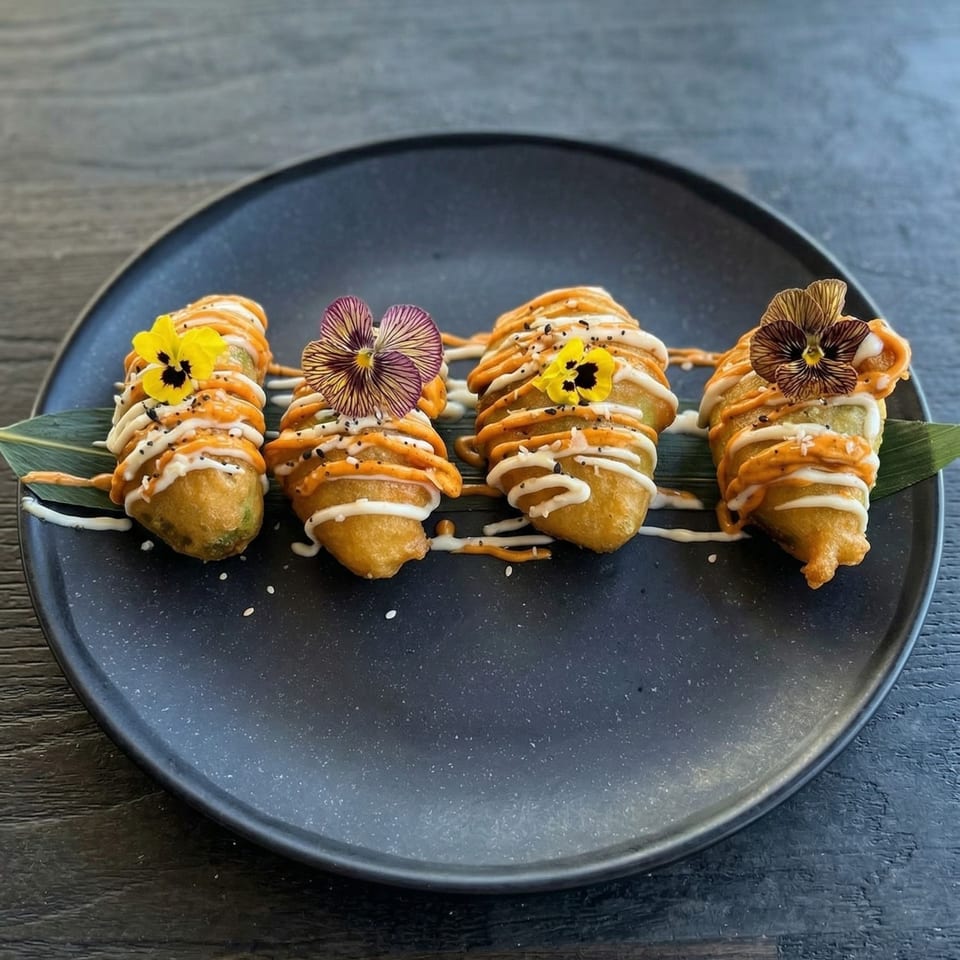 Four fried appetizers drizzled with orange and white sauces, garnished with edible flowers on a dark plate.