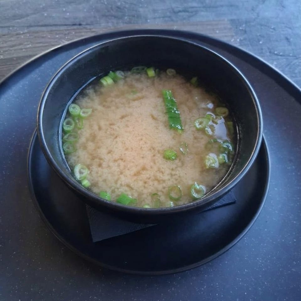 A black bowl of miso soup with green onions and tofu, served on a dark plate.