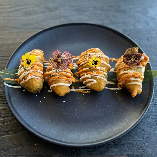 Four fried appetizers drizzled with orange and white sauces, garnished with edible flowers on a dark plate.